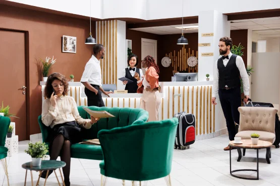 Voyageurs et personnel dans un hall d’hôtel avec comptoir d’accueil et fauteuils verts.